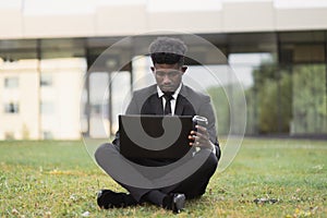 African-American man using laptop outdoors, sitting on the grass near office