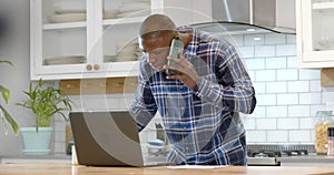 African american man using laptop and talking on smartphone in kitchen at home, slow motion