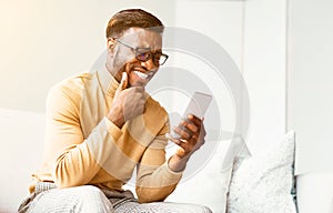African American Man Using Smatphone Texting And Laughing Sitting Indoor