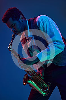 African-American man virtuoso, solo musician playing jazz melodies on saxophone against gradient blue background in