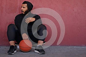 African-American man wearing a black hoodie sitting on a skateboard with a basketball, yawning and looking sideways