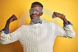 African american man wearing denim shirt and white sweater over isolated yellow background showing arms muscles smiling proud