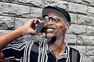 African american man, wearing hat and glasses, talking on mobile phone, standing with his back to gray brick wall