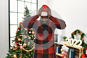 African american man wearing santa claus hat standing by christmas tree suffering from headache desperate and stressed because