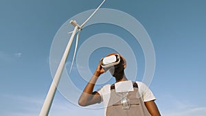 African american man wearing VR headset while standing at rural area with huge windmills around. Low angle view. New