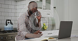African American man working at home in kitchen talking on smartphone and using laptop