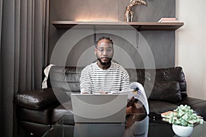 African American man working with laptop computer remote while sitting at sofa in living room. Black guy do freelance