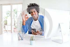 African American man working using smartphone and computer doing ok sign with fingers, excellent symbol