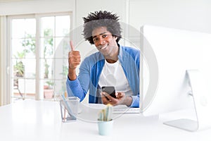 African American man working using smartphone and computer happy with big smile doing ok sign, thumb up with fingers, excellent