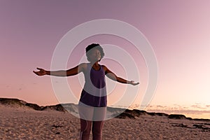 African american mature woman with afro hair closing eyes and stretching arms on beach at sunset