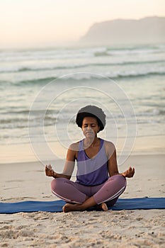 African american mature woman with afro hair meditating while sitting on mat against seascape