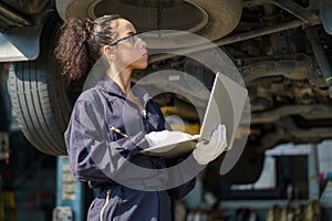 african american mechanic woman using laptop computer checking list to undercarriage of car in workshop at auto car repair service