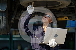 african american mechanic woman using laptop computer checking list to undercarriage of car in workshop at auto car repair service