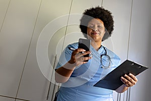 African american mid adult female nurse using smart phone while holding clipboard in hospital