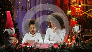African American mom and little daughter reading a letter written for Santa Claus. Woman and girl sitting near Christmas