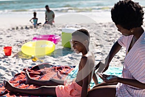 African american mother applying sunscreen on shirtless son's body while sitting on beach