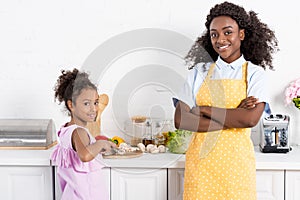 african american mother with crossed arms and daughter cutting vegetables