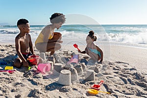 African american mother crouching by children making sandcastles at beach on sunny day