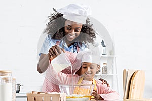 african american mother and daughter in chef hats pouring milk in bowl with ingredientes photo