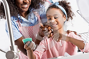 african american mother and daughter having fun while washing dishes with sponge