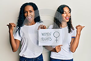 African american mother and daughter holding women rights banner pointing thumb up to the side smiling happy with open mouth