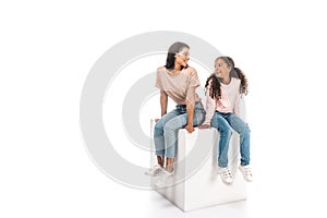 African american mother and daughter looking at each other while sitting on white cube on white background