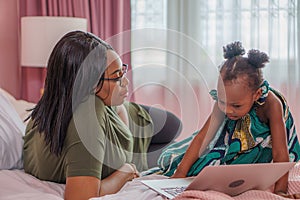 African American mother and daughter lying on bed using  laptop computer together in bedroom at home . happy black mom and girl