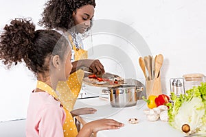 african american mother and daughter putting ingredientes in saucepan photo
