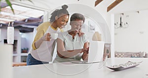 African american mother and daughter smiling while using laptop together at home