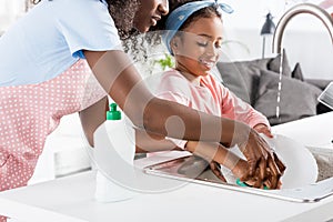 african american mother and daughter washing dishes with detergent