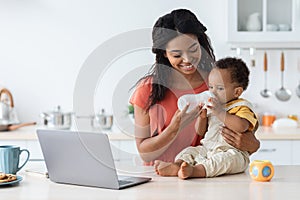 African American Mother Giving Baby Water Bottle To Infant Son In Kitchen