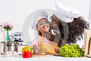 african american mother kissing her daughter in chef hat while cooking