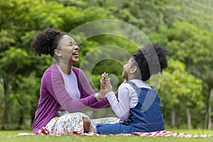 African American mother is laughing while playing patty cake with her young daughter while having a summer picnic in the public