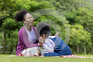 African American mother is laughing while playing patty cake with her young daughter while having a summer picnic in the public