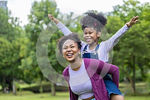 African American mother is playing piggyback riding with her young daughter while having a summer picnic in the public park for