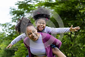 African American mother is playing piggyback riding with her young daughter while having a summer picnic in the public park for