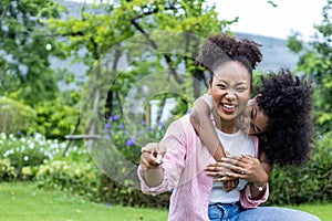 African American mother is playing piggyback riding with her young daughter while having a summer picnic in the public park for