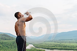 african american muscular man drinking water after sports training. Breathtaking green mountain landscape on