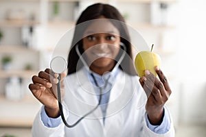 African american nutritionist showing organic green apple and using stethoscope, closeup