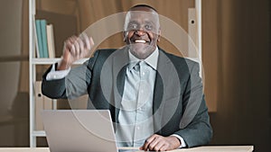 African American old senior businessman boss worker man sitting at table in office with laptop looking at camera waving