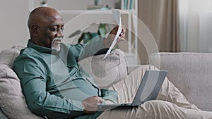 African American old senior mature man waving paper document suffering from summer heat at home working with laptop