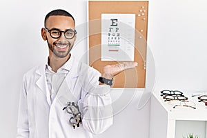 African american optician man standing by eyesight test smiling cheerful presenting and pointing with palm of hand looking at the