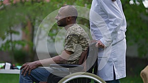 African American paralyzed man greeting unrecognizable Caucasian nurse pushing wheelchair leaving. Young caregiver