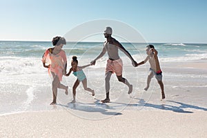 African american parents and children holding hands while running together at beach on sunny day