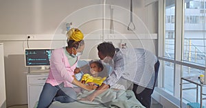 African-American parents in safety mask visit girl patient lying in medical bed in ward kids hospital