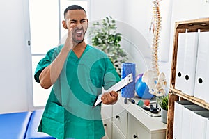 African american physiotherapist man working at pain recovery clinic shouting and screaming loud to side with hand on mouth