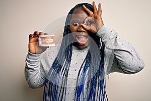 African american plus size woman with braids holding plastic teeth over white background with happy face smiling doing ok sign