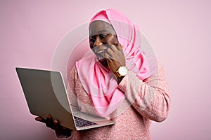 African american plus size woman wearing muslim hijab using laptop over pink background laughing and embarrassed giggle covering