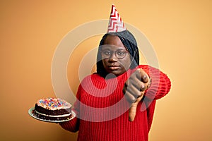 African american plus size woman wearinng birthday hat holding cake over yellow background with angry face, negative sign showing