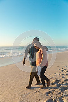 African american retired senior couple walking together at beach against blue sky with copy space